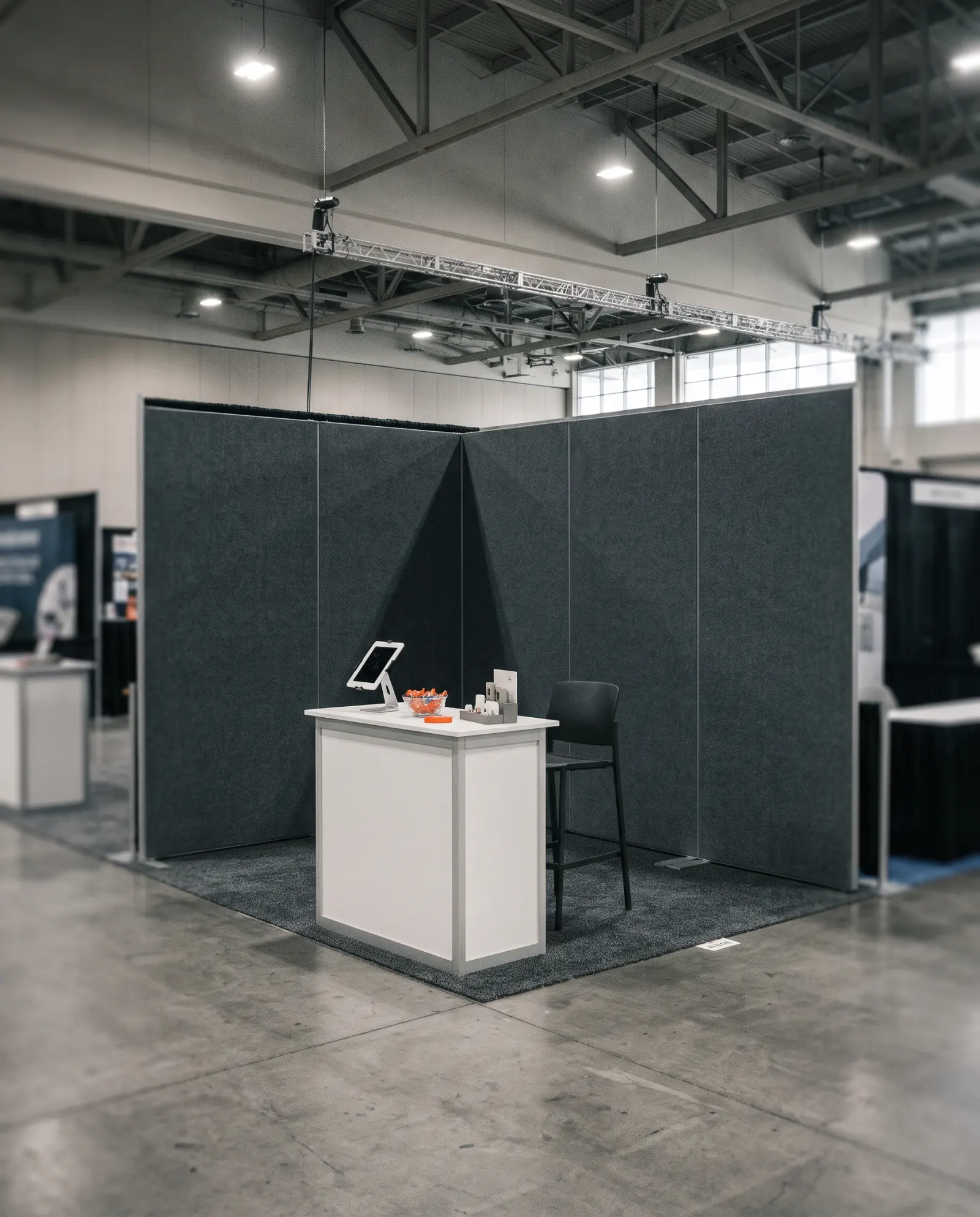 A small inline trade show booth before opening: charcoal fabric walls, a white counter with a tablet and a bowl of candy, a tall stool on grey carpet